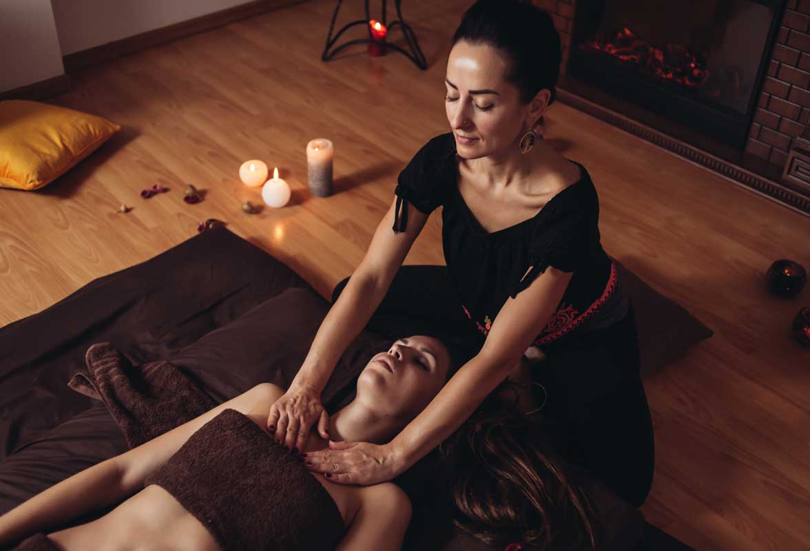 Séance de massage dans une atmosphère relaxante avec bougies et lumière tamisée. Une femme masse le haut du corps d'une cliente allongée, soulignant le bien-être et la détente