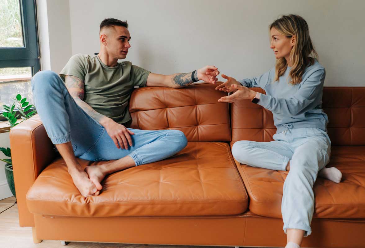 Un couple assis sur un canapé en cuir marron discute. L'homme et la femme sont en tenues décontractées, engagés dans une conversation ou un désaccord dans un salon lumineux