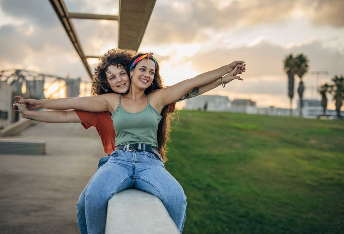 Les styles d’attachement : comprendre ta façon d’aimer Deux jeunes femmes sont assises sur un muret en béton, se tenant mutuellement avec les bras écartés, comme si elles volaient, dans une pose joyeuse et insouciante.