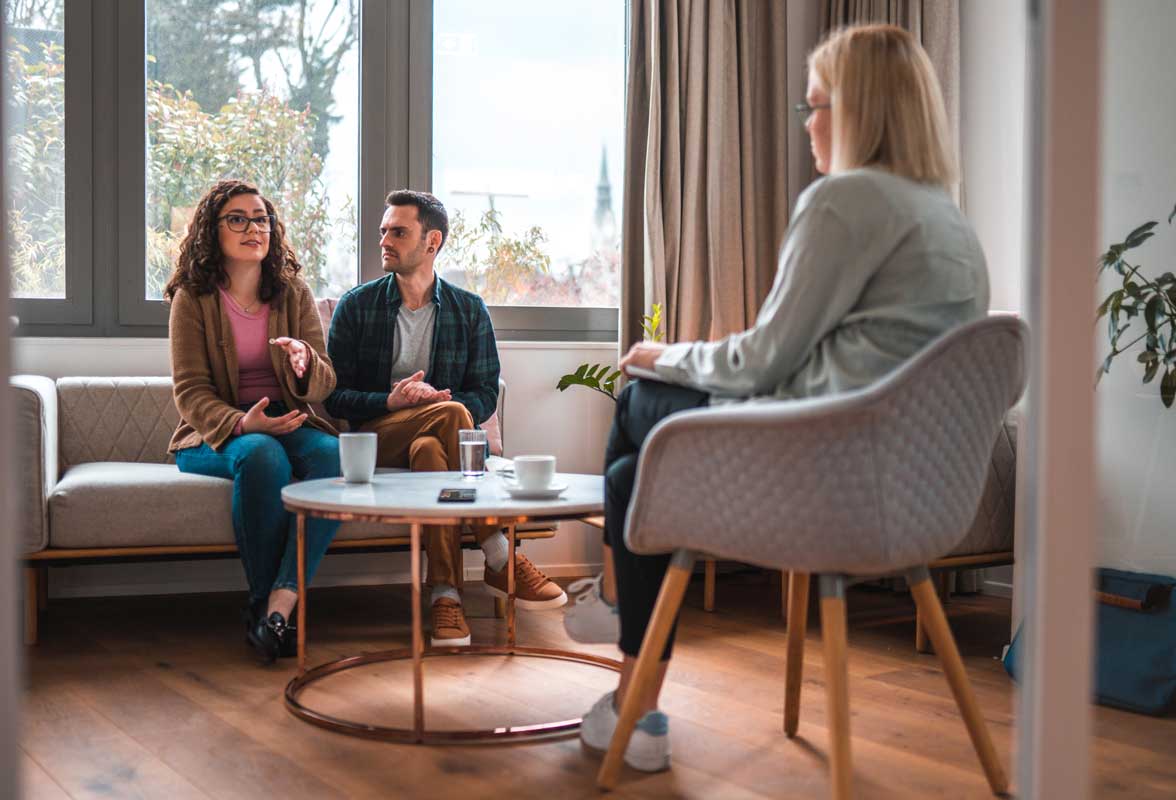 Les styles d’attachement : comprendre ta façon d’aimer Séance de thérapie de couple. Un homme et une femme sont assis sur un canapé, discutant avec une thérapeute assise en face, dans un bureau lumineux et moderne