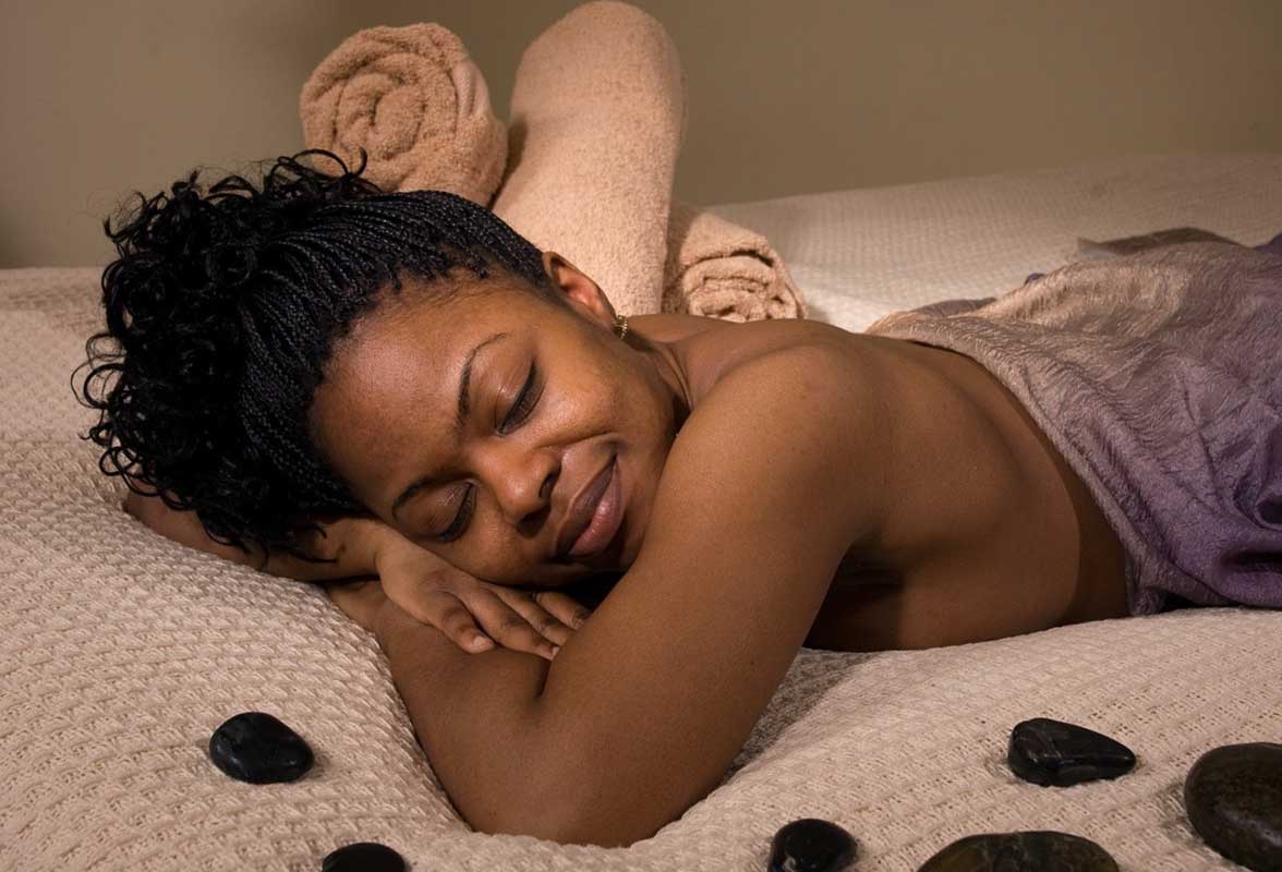 Femme souriante et détendue, les yeux fermés, allongée sur le ventre pour un massage bien-être. Elle est entourée de quelques pierres de massage sombres sur le lit.