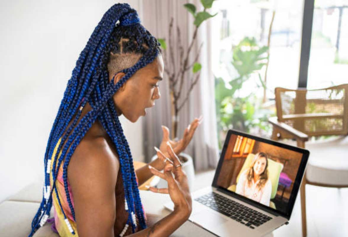 Séance de consultation en ligne. Une personne avec de longues tresses bleues parle avec des gestes expressifs à son écran d'ordinateur, où l'on voit une thérapeute en sexothérapie souriante.